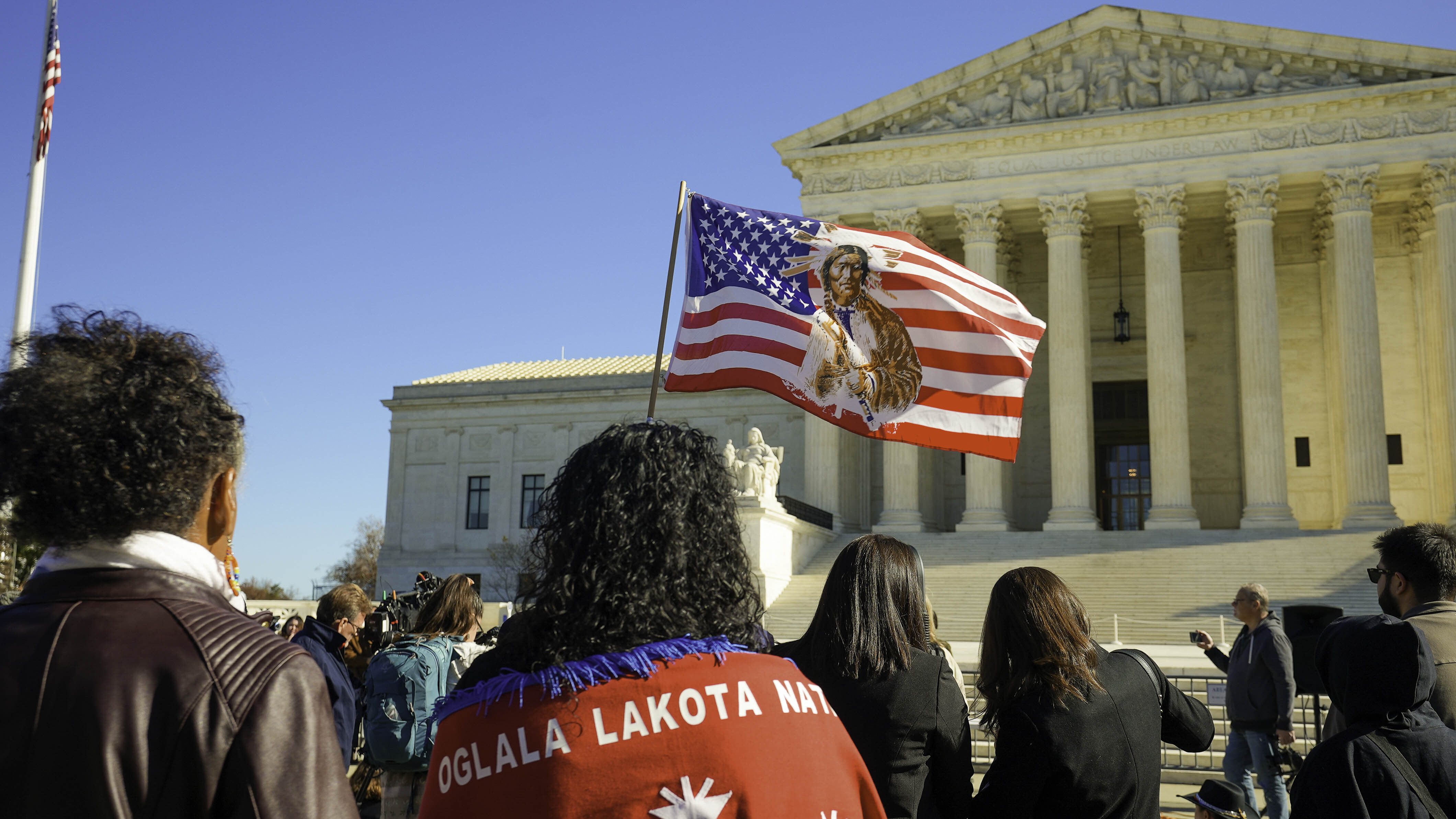 An image shows Native Americans and media gathered outside the Supreme Court Building in Washington, D.C. waiting for a decision. One Native American holds a powerful flag featuring Sitting Bull's image on an American flag.