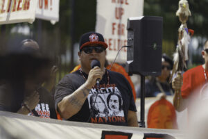 An image of a Native American man wearing a snapback hat from the American Indian Movement and speaking through a microphone during a protest.