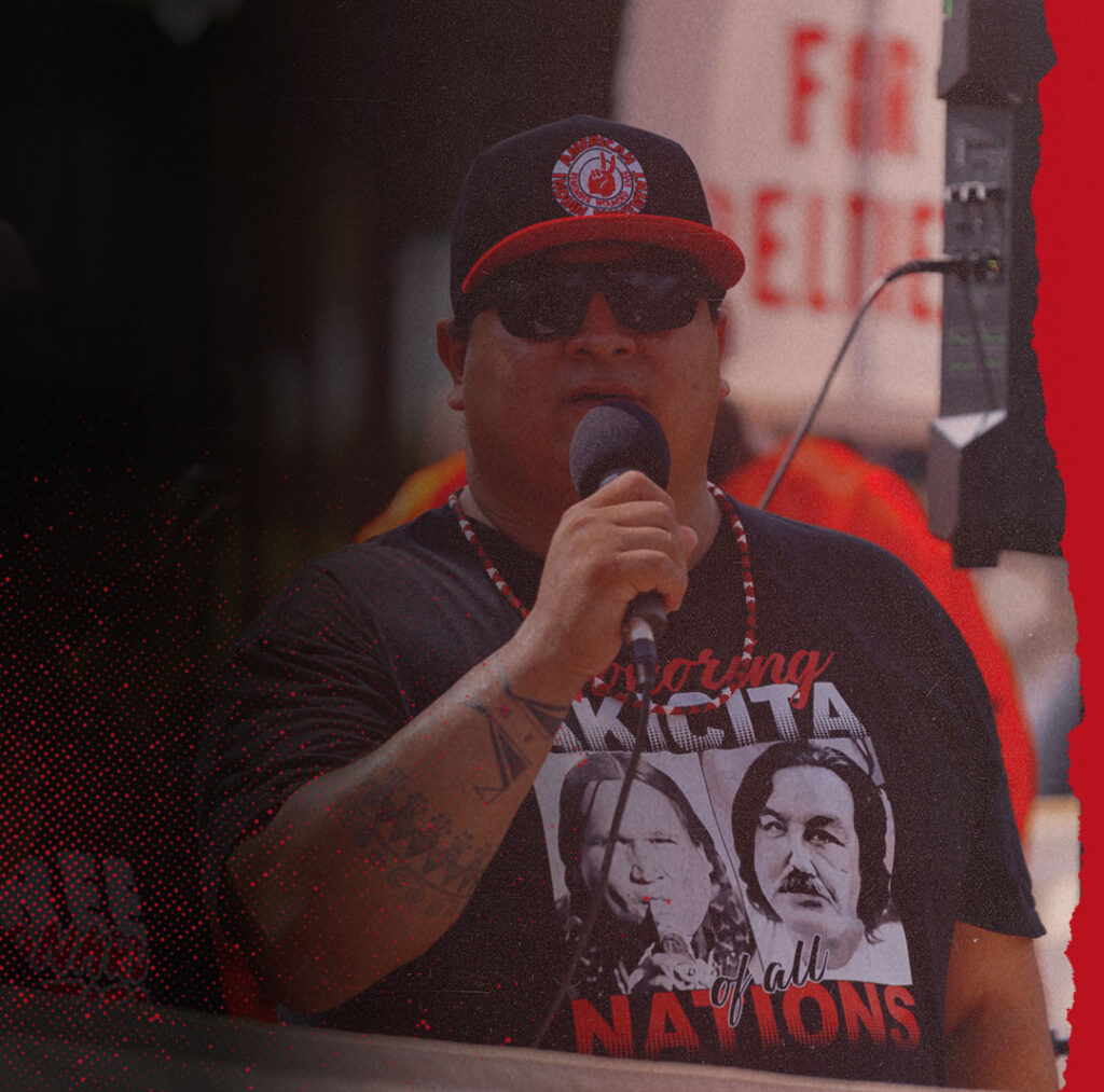 An image of a Native American man wearing a snapback hat from the American Indian Movement and speaking through a microphone during a protest.