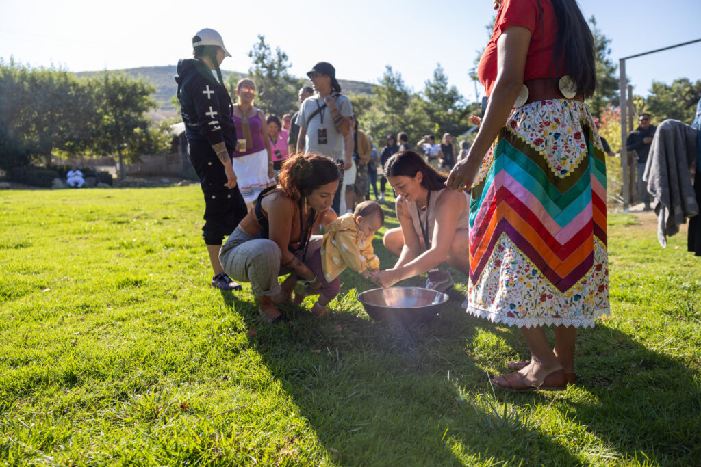 Two Indigenous women hold a baby girl over a metal bowl of water to wash her hands while a woman in a colorful ribbon skirt watches. 