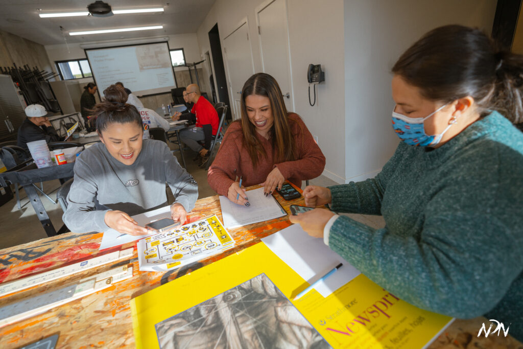 Three women sit around a table with pens and paper, two of them are laughing together and one wears a blue face mask. 
