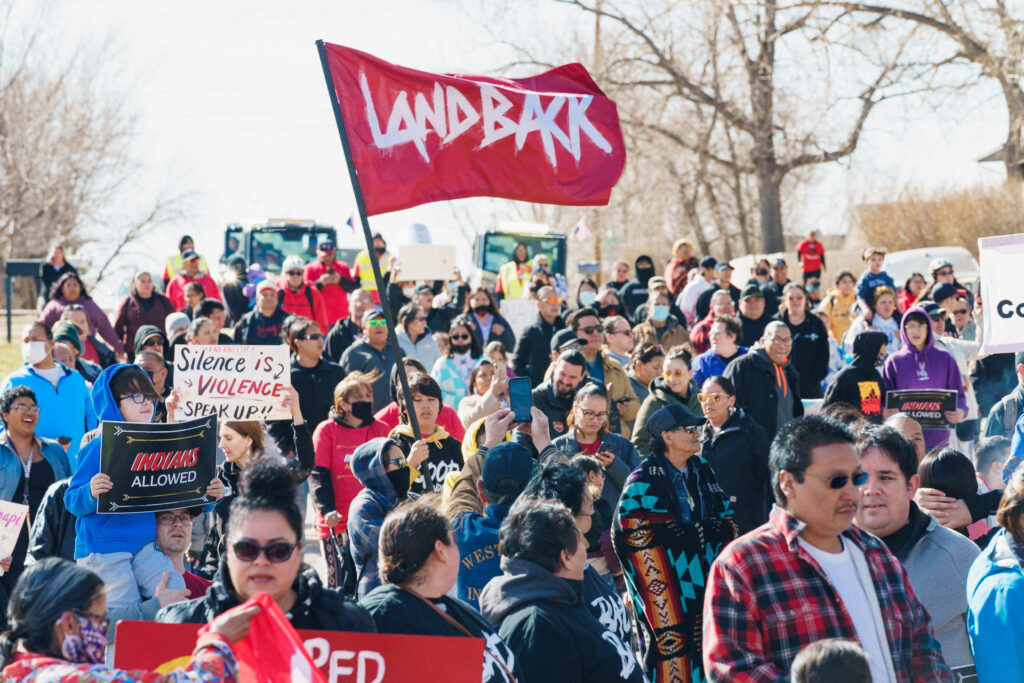 A crowd marches through the street in winter. A young man carries a bright red flag that says LANDBACK and other carry signs that say 'Silence is Violence, Speak Up!!' and 'Indians Allowed'. 