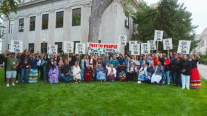 A group of Indigenous people gather in front of a building holding signs with their fists held high in the air.