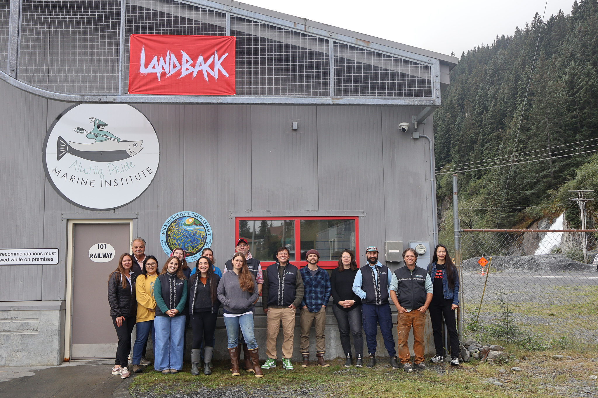 Indigenous people stand in front of a building with a logo of a person canoeing that read Alutiiq Pride Marine Institute. A red flag that says LANDBACK hangs from the roof.