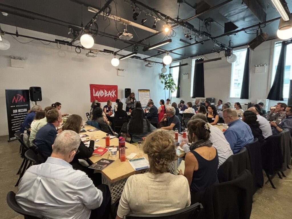 Tables are arranged in a large square and every seat is filled. At the front of the room there is a bright red LANDBACK sign and to the left a NDN Collective banner that has the mission printed on it. In front of the room a woman speaks on the mic.