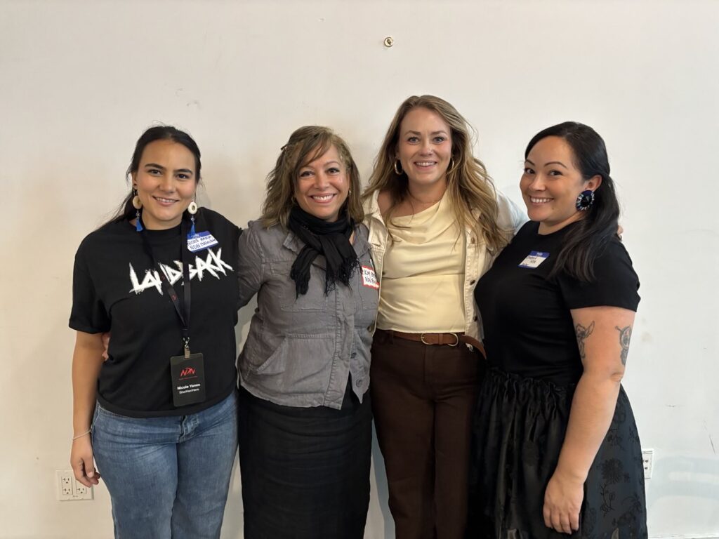 Four women stand arm and arm against a while wall smiling. The woman to the left wears a black LANDBACK shirt, to her right stands a woman in a gray cargo jacket, next to her is a woman in a yellow top, and the woman on the far right stands in an all black dress with tattoos on her arm.