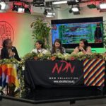 four women sit at a table. in the background there is a bright red light illuminating 'The Peoples Forum', a TV and green plants. The women sit at a table with a cloth that reads NDN Collective.