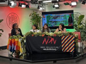 four women sit at a table. in the background there is a bright red light illuminating 'The Peoples Forum', a TV and green plants. The women sit at a table with a cloth that reads NDN Collective.