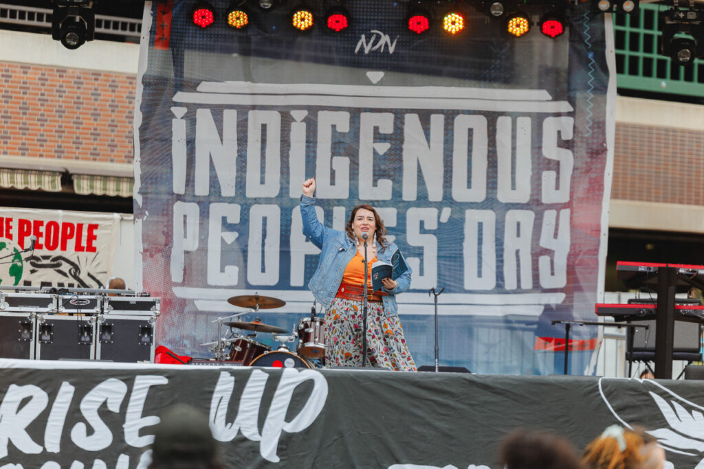 A woman stands on stage in a denim jacket and flowered skirt with her right fist raised and her left hand holds a book.