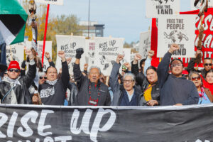 People stand at the front of a large crowd. A middle-aged man stands next to an elderly man and woman with their fists raised.