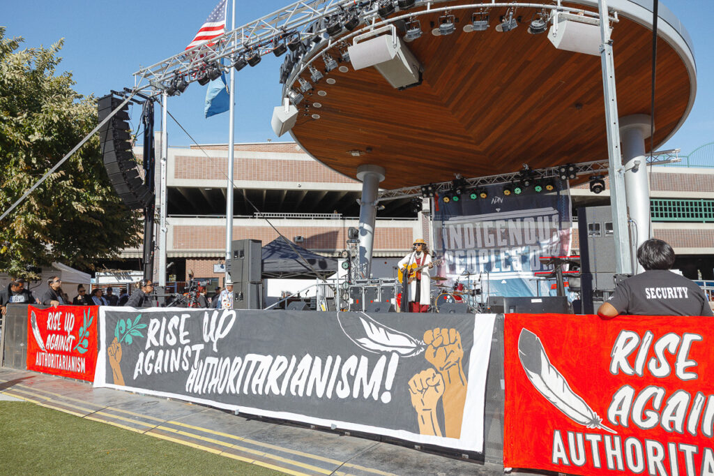 A woman in a white coat is standing on stage in front of a black and white banner that reads RISE UP AGAINST AUTHORITARIANISM.