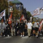 A group of people march along a road in a town to celebrate Indigenous Peoples day. Some carry tribal flags or LANDBACK flags.