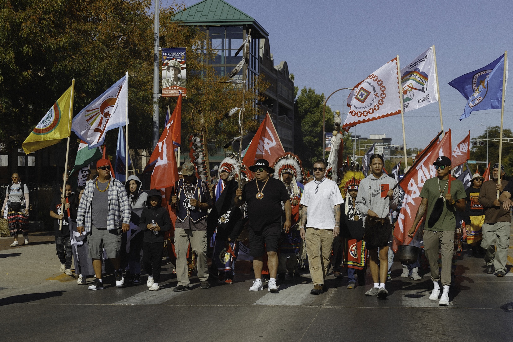 A group of people march along a road in a town to celebrate Indigenous Peoples day. Some carry tribal flags or LANDBACK flags.