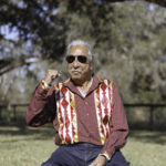 Leonard Peltier sits in a chair outside with his fist up in celebration.
