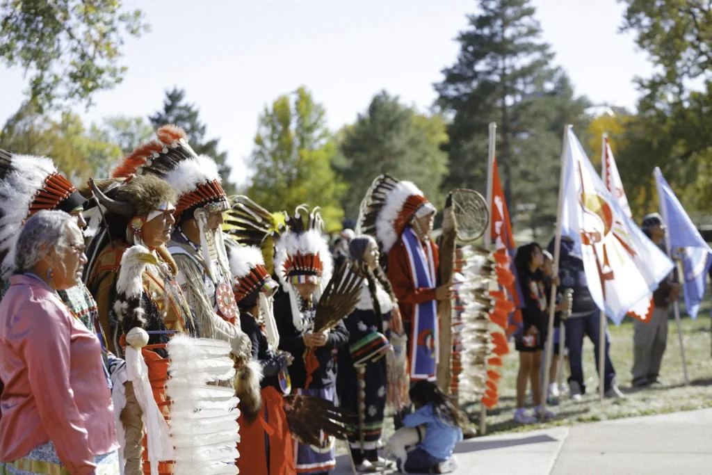 Tribal leaders gathered outdoors in regalia with flags at Indigenous Peoples’ Day in 2024.
