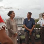 Three people lean against a low fence in conversation. They are dressed in business wear and large white regal buildings are in the background.